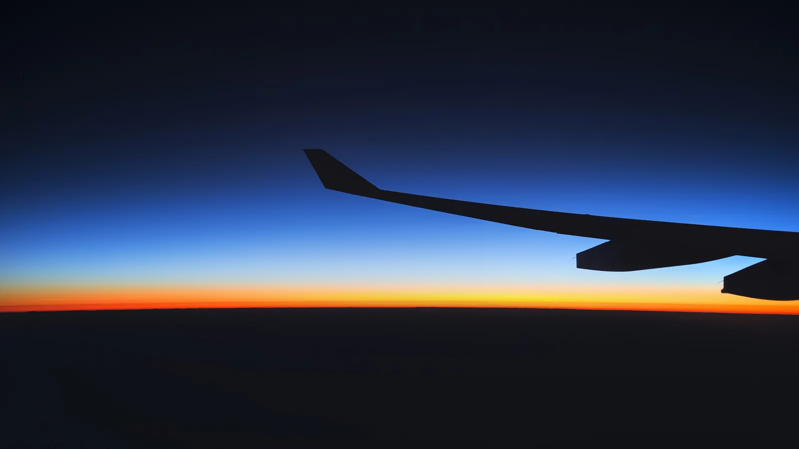 A dark airplane wing silhouetted against a twilight sky, with an orange glow along the horizon.