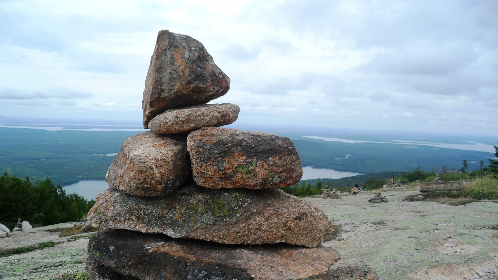 A cairn of flat granite rocks stacked on a mountaintop, with lakes and forest far below.