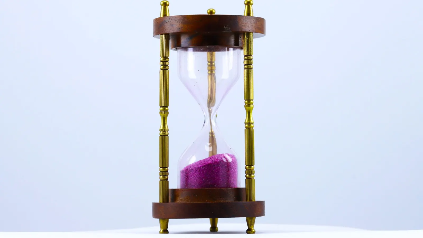 An hourglass with wooden ends and brass rods holds pink sand against a pale background.