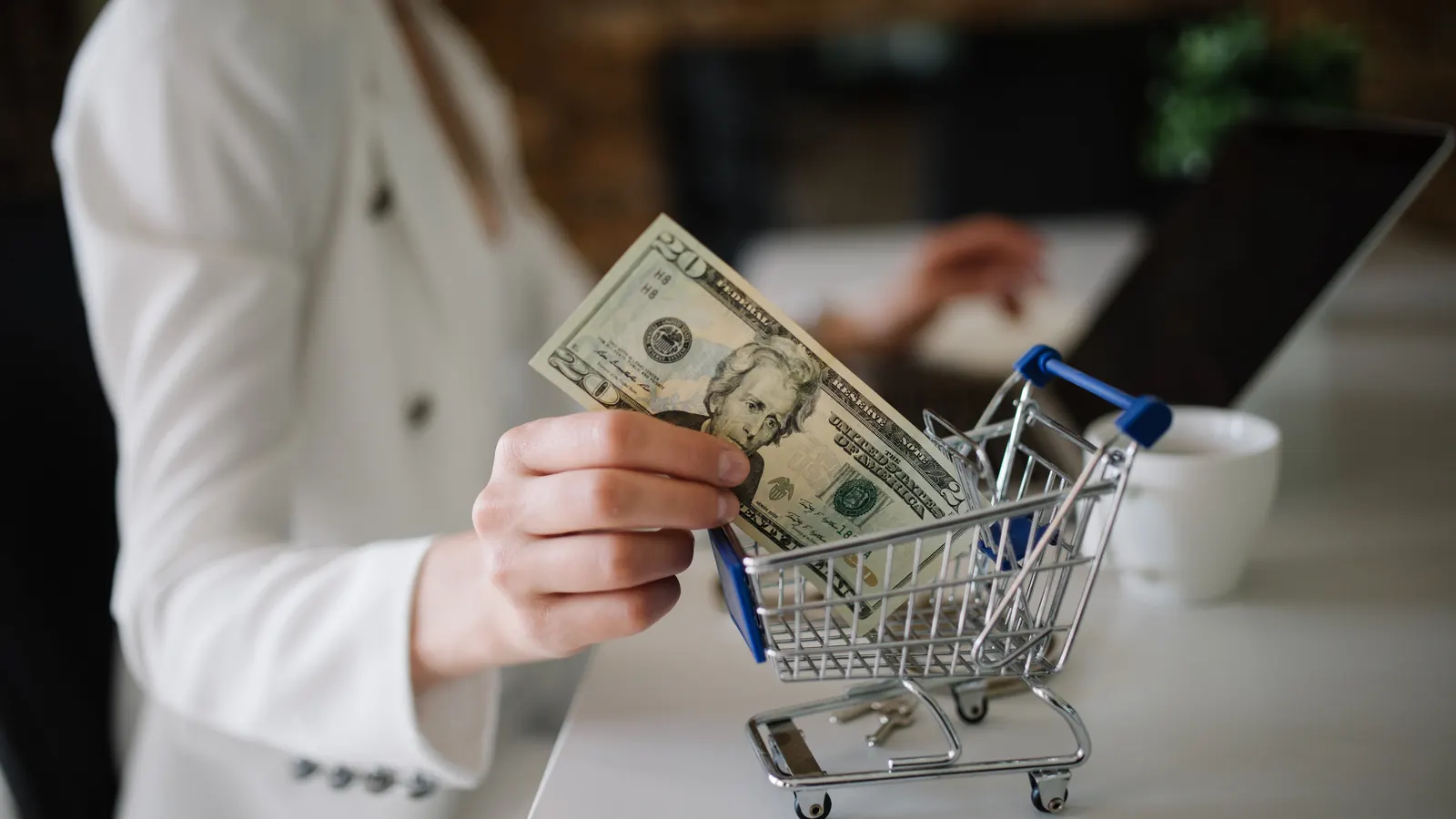 A hand holds cash over a small shopping cart on a table beside a laptop.