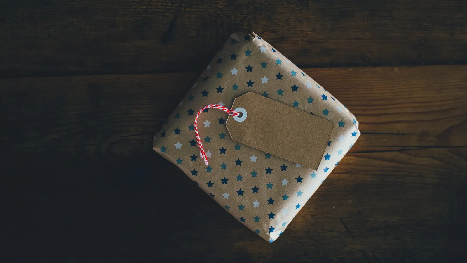 A star-printed gift wrapped in brown paper, with a blank kraft tag on red and white twine, on dark wood