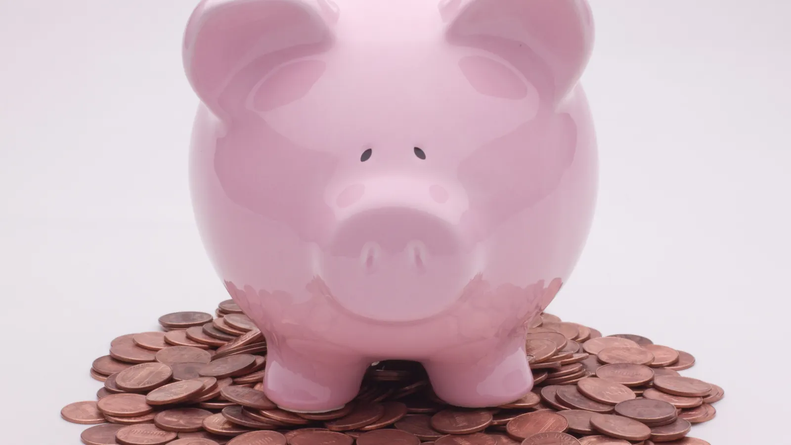 A pink ceramic piggy bank standing on a heap of copper pennies against a pale background
