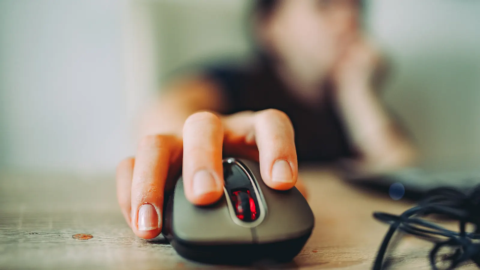 A hand rests on a computer mouse on a wooden desk, with a blurred background.