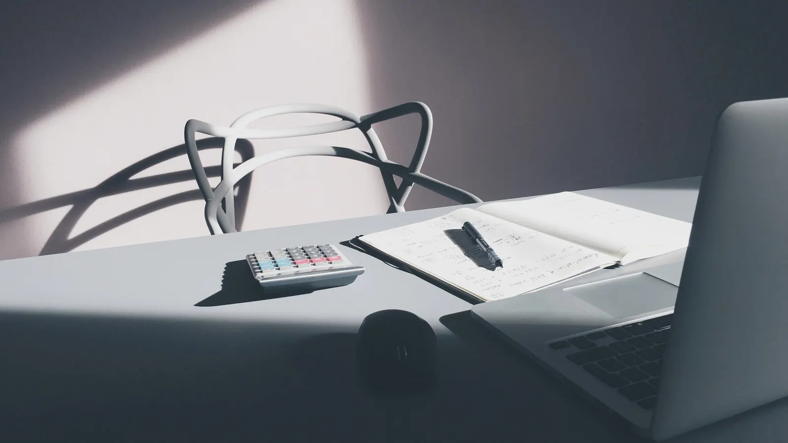 A white desk calculator beside an open notebook with a pen, and a laptop at the edge of the desk.