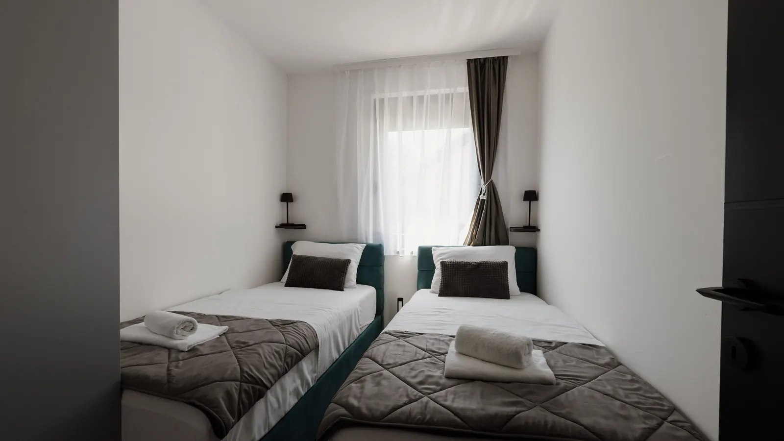 Two single beds with patterned covers and rolled towels in a bright, plain hotel room by a window.