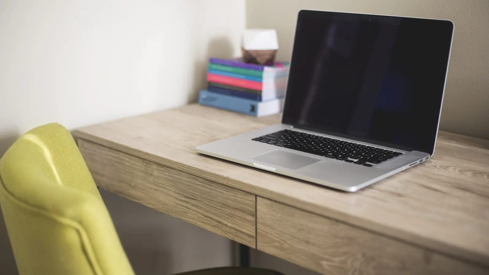 An open laptop on a light wooden desk beside a stack of books and a yellow chair