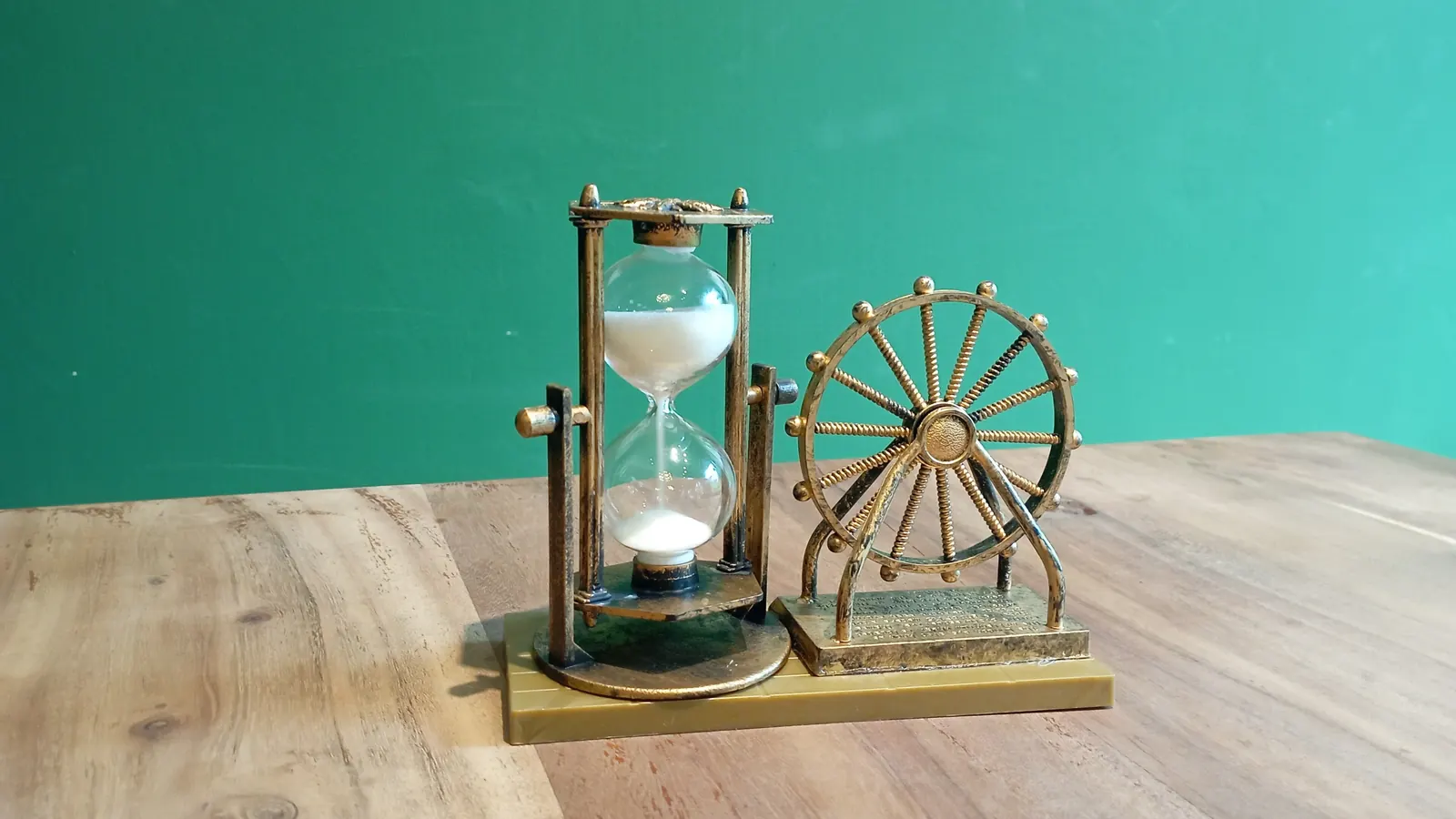 A brass hourglass in a small frame beside a spoked brass wheel, on a wooden table