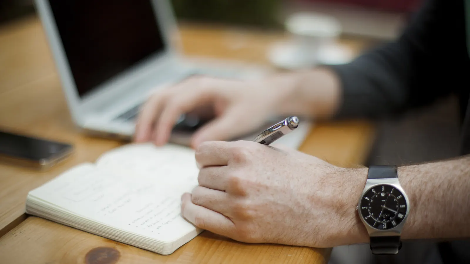 A hand writing in a small notebook with a pen, a laptop and phone nearby, a wristwatch on the wrist