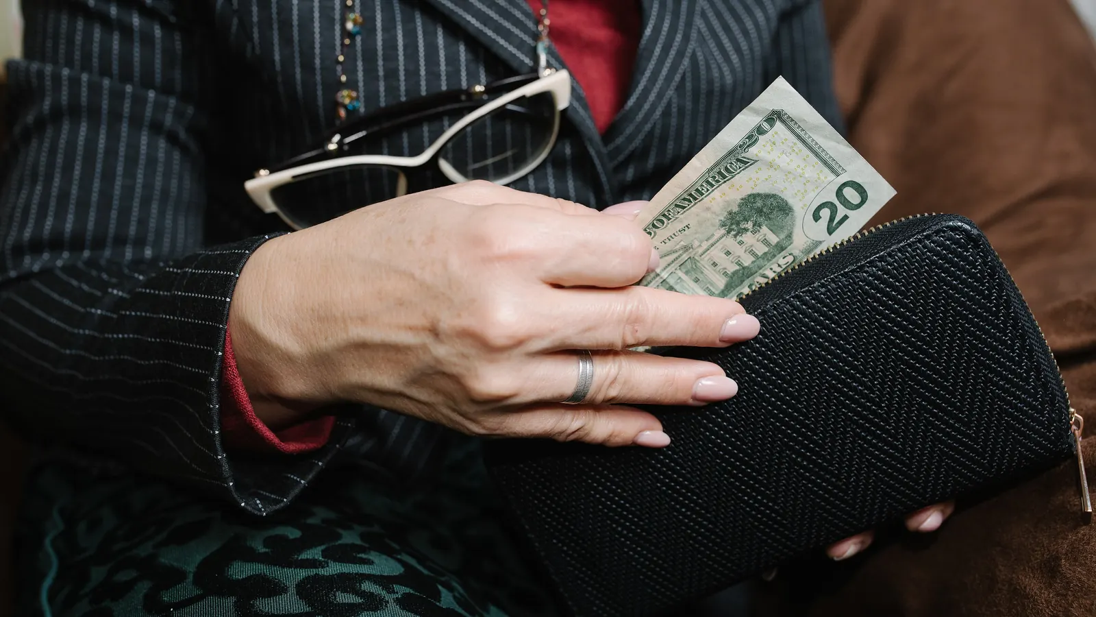 A hand pulls a twenty-dollar bill out of a black wallet resting on someone's lap