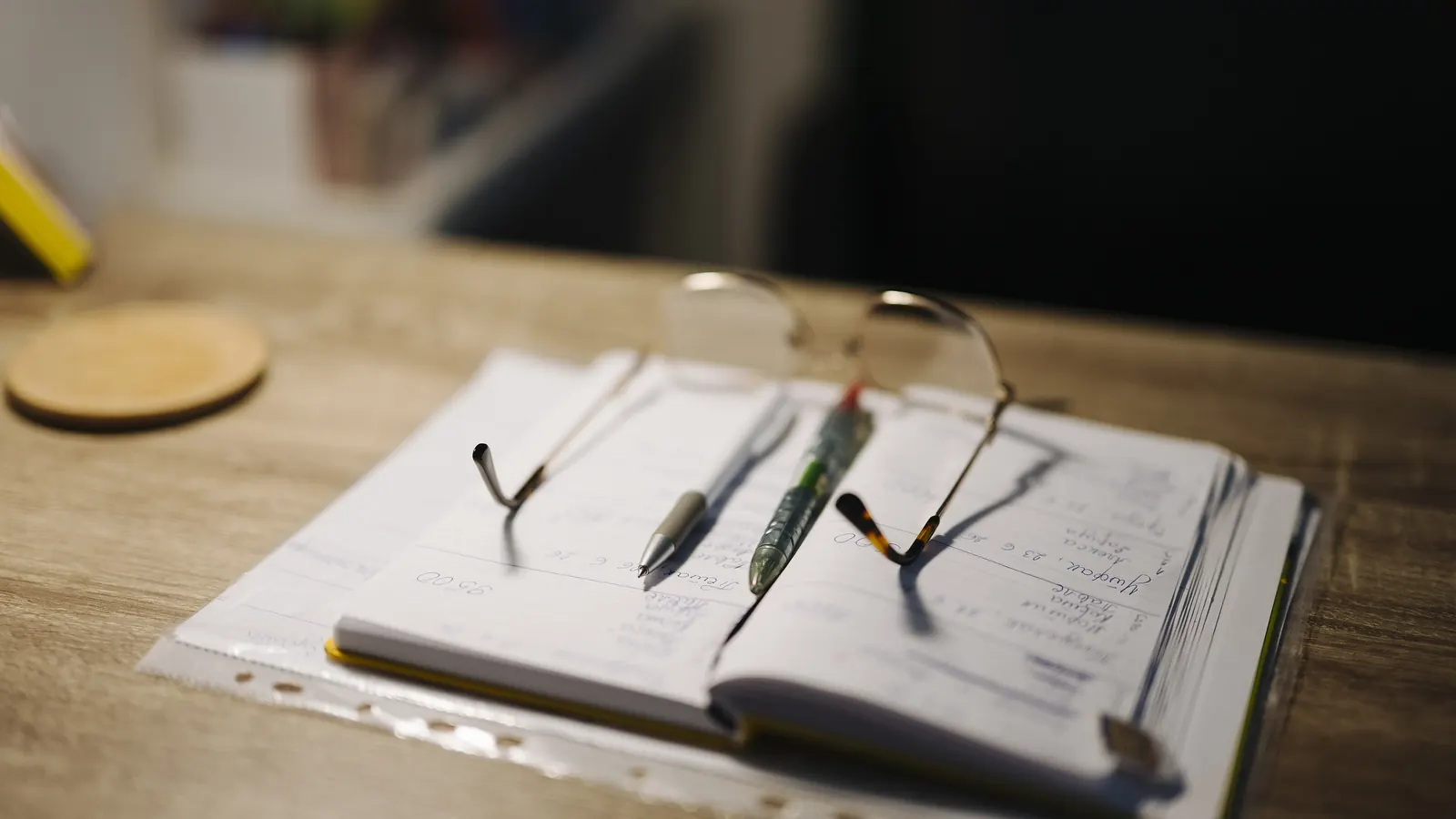 An open notebook with handwritten notes, two pens and a pair of glasses on a wooden desk.