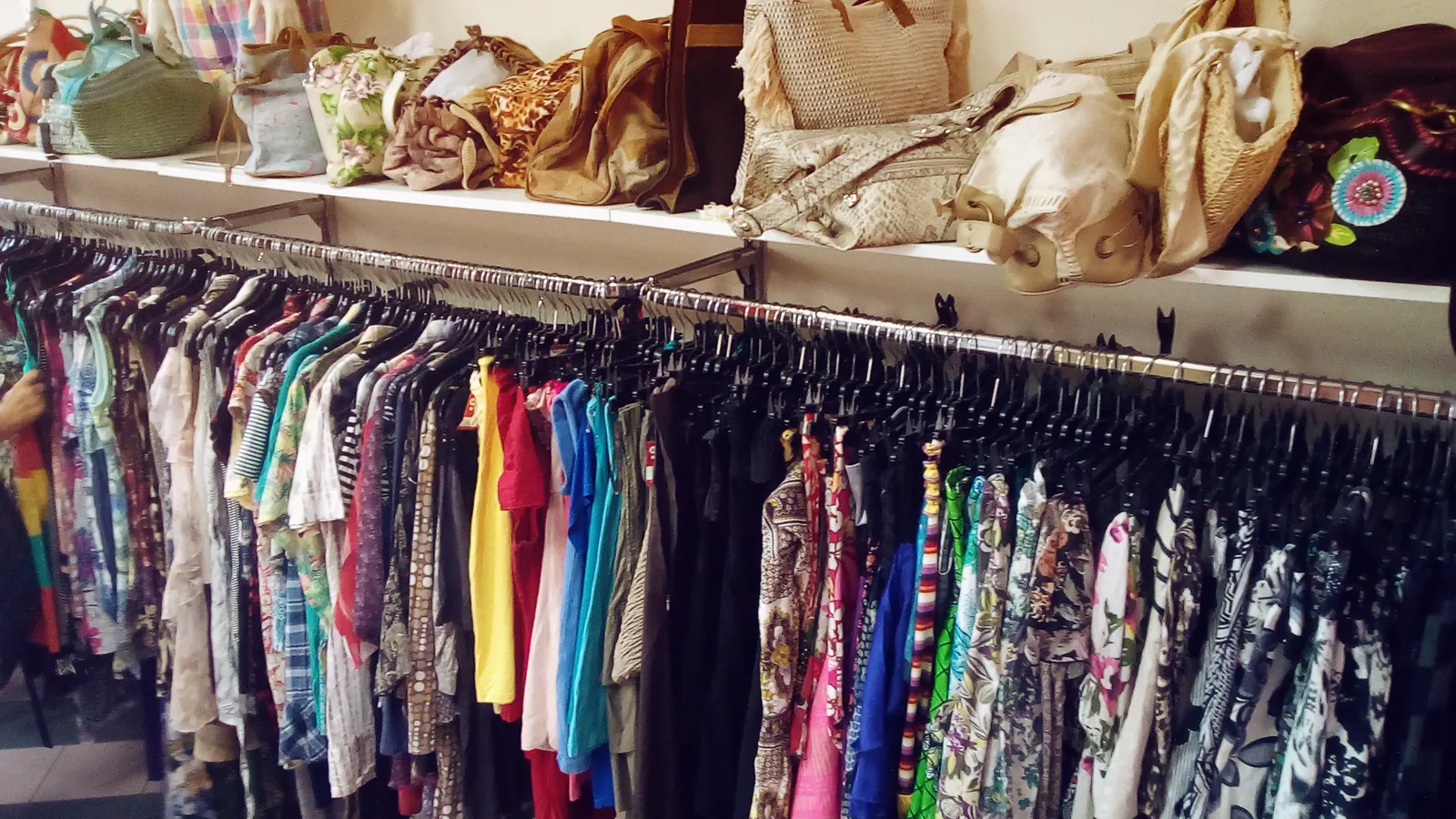 Racks of colorful clothes on hangers, with handbags on a shelf above, inside a shop.