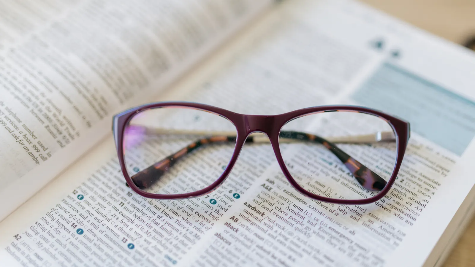 Burgundy-framed reading glasses resting on an open dictionary page with printed entries